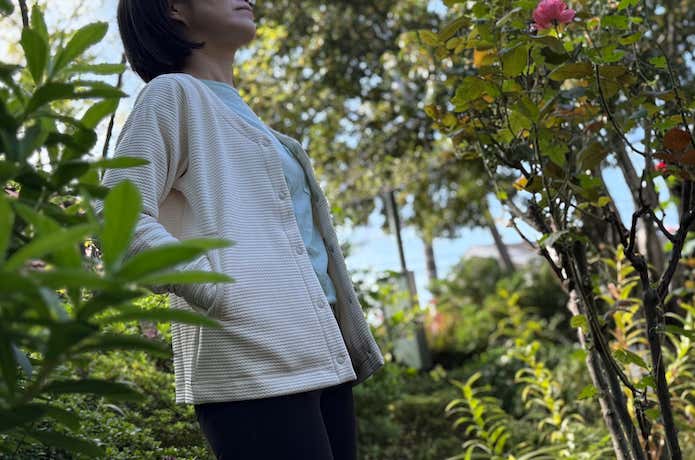 リップルニットカーディガンを羽織って草木の間に立つ人